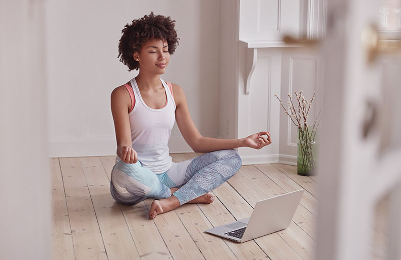 Woman meditating cross-legged on wooden floor with laptop nearby.