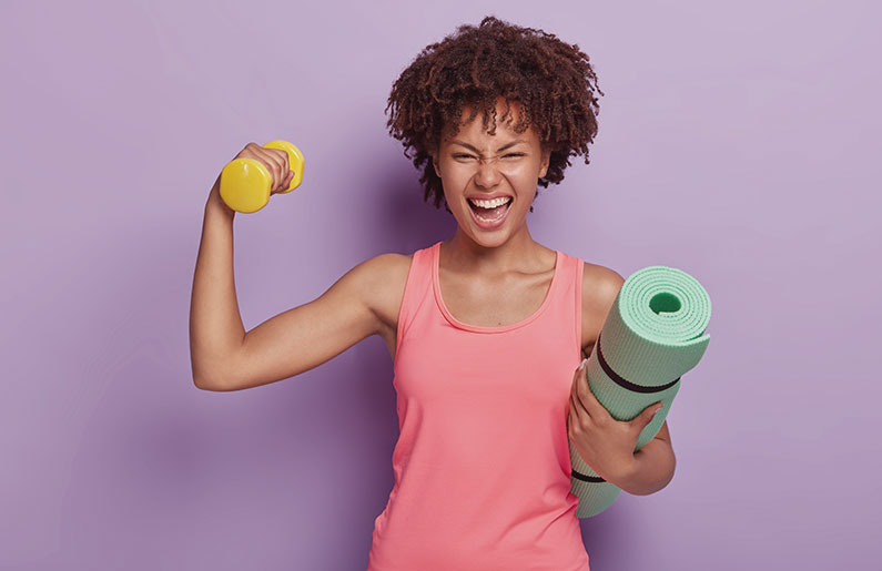 Woman flexes arm with dumbbell and yoga mat, showing fitness enthusiasm.